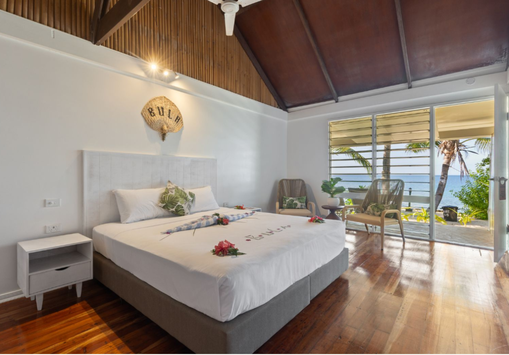 Interior of a Beachcomber Island bure with a neatly made bed and open windows framing the turquoise ocean.