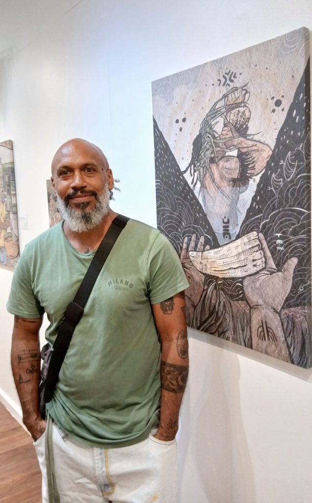ALT TEXT: Ropate Kama standing beside one of his artworks from Vulaiwerewere: Celebrating the Moons of the iTaukei Calendar at the Epeli Hau’ofa Art Gallery, Oceania Centre at USP in Suva. EXPLORE Fiji photo. 