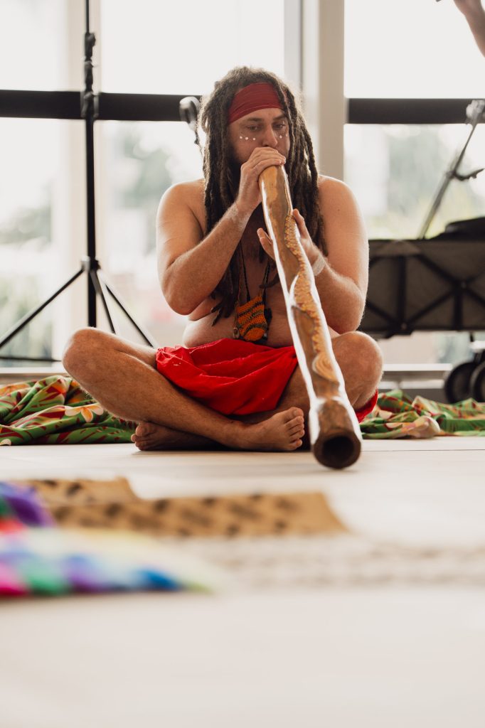 An Aboriginal Australian man in traditional ceremonial attire playing a wooden didgeridoo wind instrument at the Gold Coast Airport terminal event.