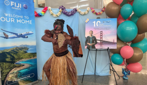 A Fijian warrior in traditional attire performing a cultural dance move in front of Fiji Airways promotional branding at the SFO airport terminal.