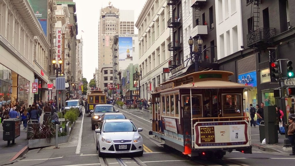 Two classic San Francisco cable cars on tracks travelling alongside a line of modern motor vehicles on a city street