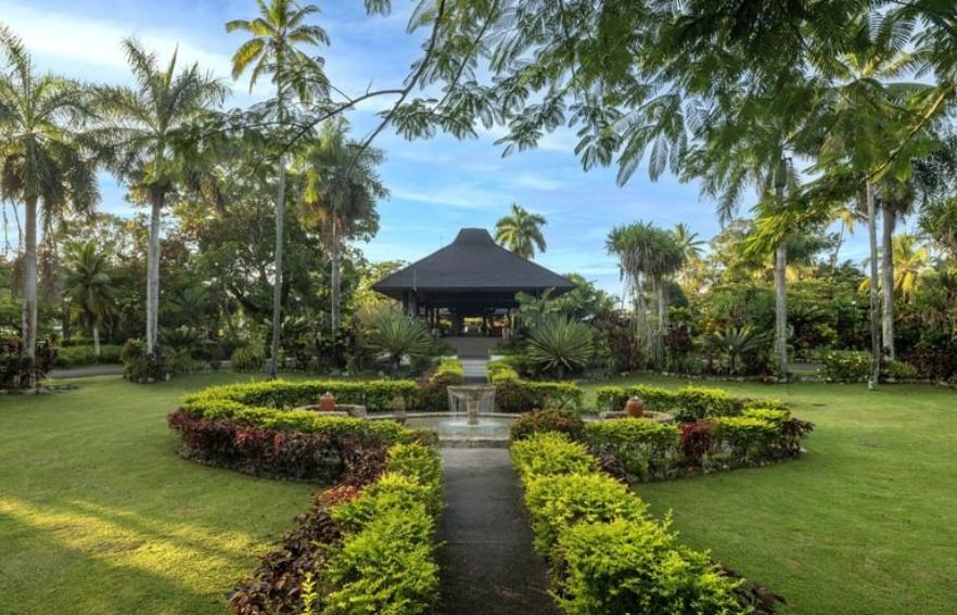 A grand thatched-roof entrance pavilion surrounded by lush, manicured tropical gardens and palm trees at a luxury resort