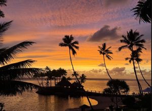 Wide panoramic shot of a vibrant sunset over a calm tropical ocean lagoon with silhouetted palm trees at dusk at The Warwick Fiji along Viti Levu's Coral Coast in Fiji