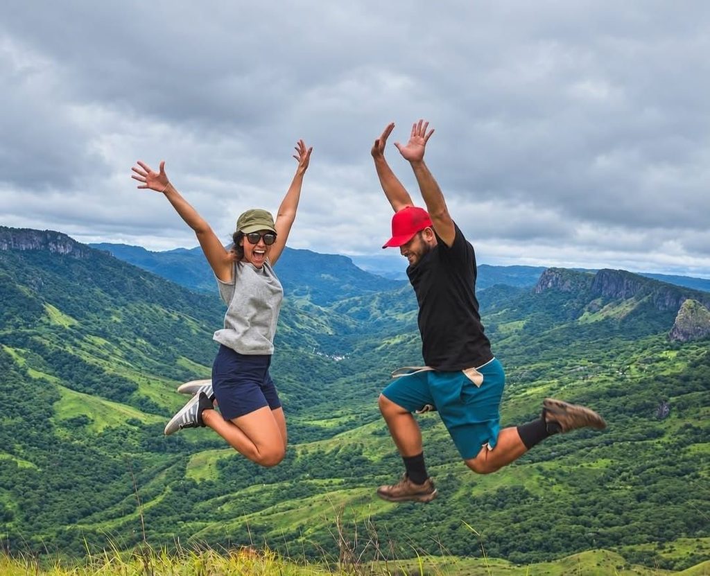 Raymond Lee (right) and a visitor jump for joy at the summit of the highest peak in the Nausori Highlands, with lush green mountain ridges and the valley towards Natasa Village and the Navosa Mountain Range in the background.