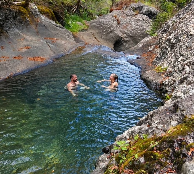 A man and woman relax in the natural pool at Vereni Falls, surrounded by lush tropical vegetation and the tranquil beauty of the forest.