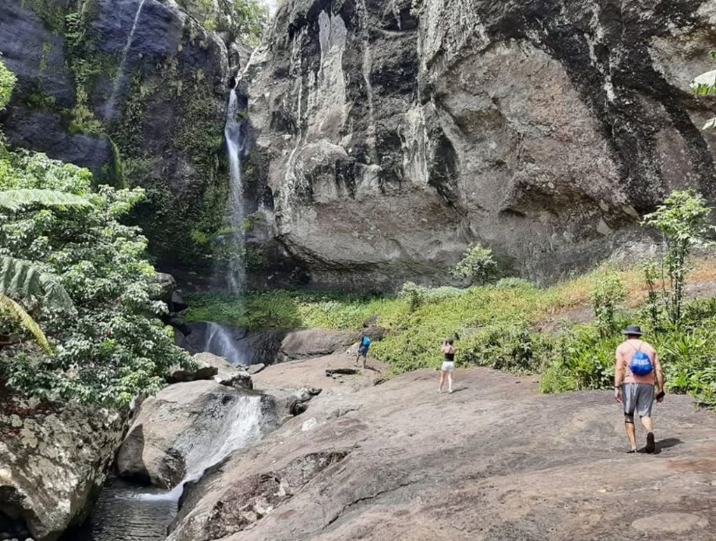 Three hikers walk across a rocky trail towards a cascading waterfall beneath a towering rock face at Tunutuni Waterfall in Koroyanitu National Heritage Park, Viti Levu, Fiji.
